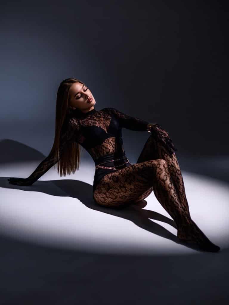 Visual Hook: Dramatic spotlighting illuminates a figure in intricate lace and fishnet,...