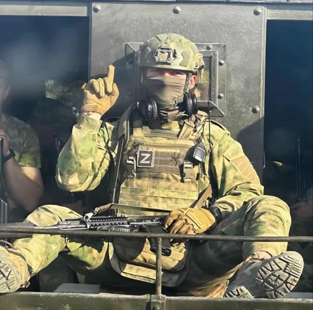 Visual Hook: A soldier in full camouflage gear and helmet, seated in a military vehicle...